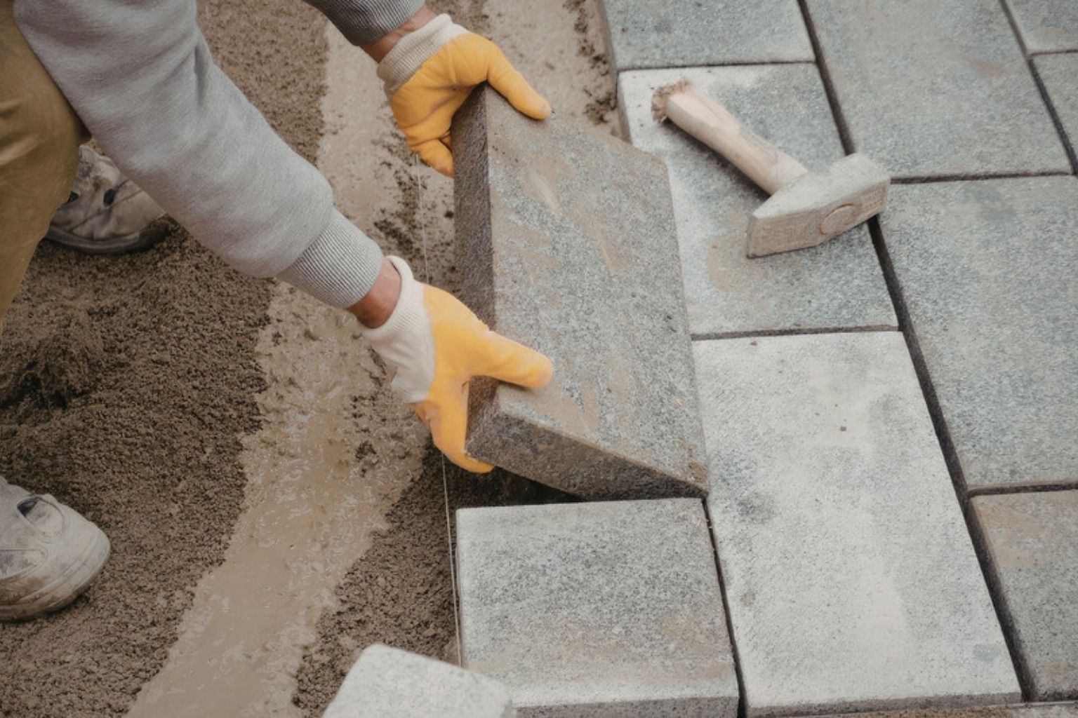 Hænder i arbejdshandsker lægger en betonflise på et sandlag med en gummihammer ved siden af